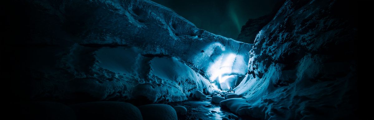 A person shining a light on the blue walls of a glacier in Iceland at night with a streak of green northern lights in the sky.